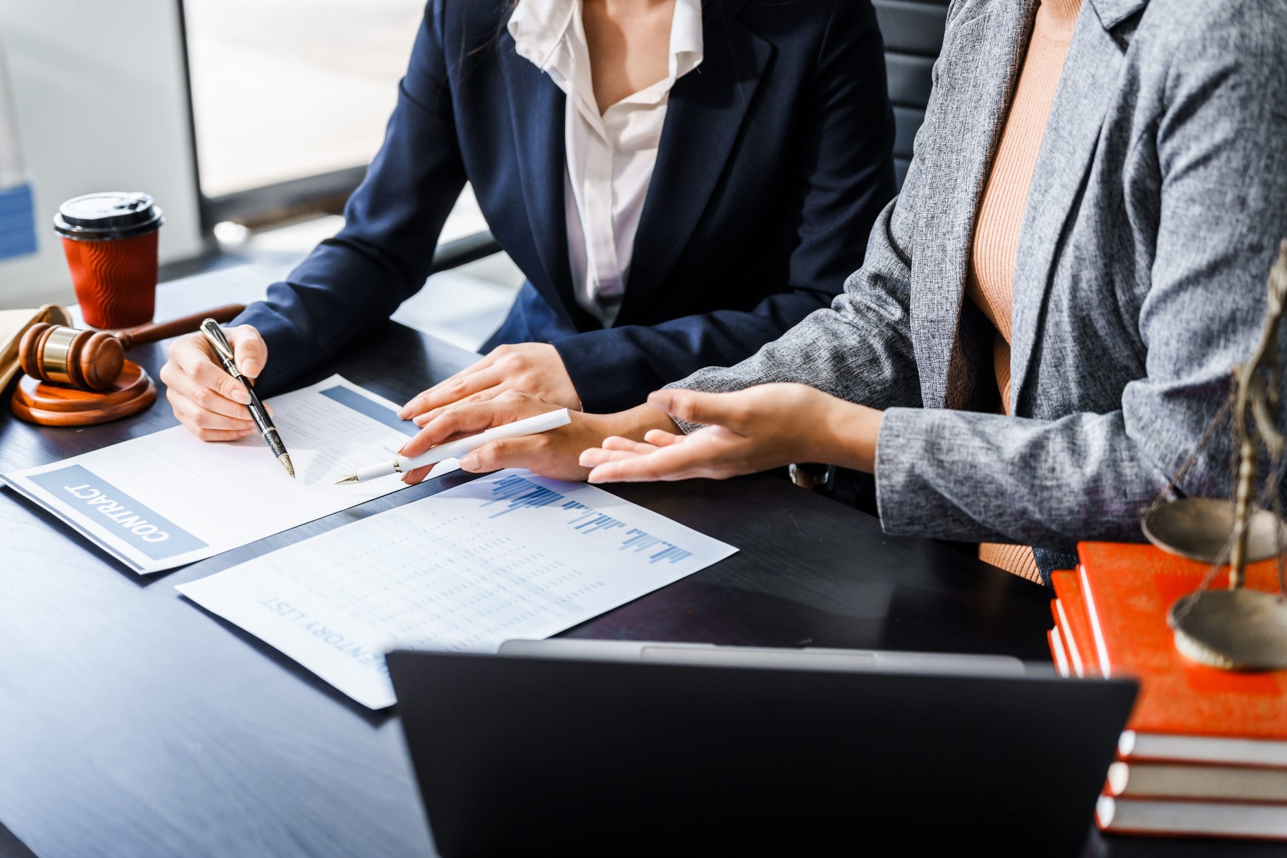 Two female lawyers in business suits meet at wooden desk, shaking hands over contract paper. wooden gavel and scales justice as discuss legal compliance, corporate law, dispute resolution.